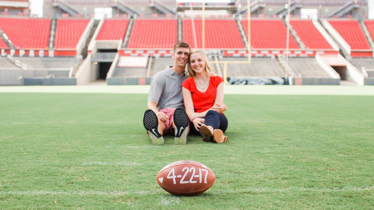 A photo of Wil Baumann and Olivia Clapp Baumann — a married couple who met at NC State — posing on the field at NC State's Carter-Finley Stadium with a football showing the date of their anniversary.