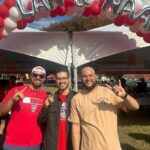 A trio of Wolfpackers throw up the Wolfie hand signal under red and white balloons at an NC State Native American Alumni Network outdoor event.