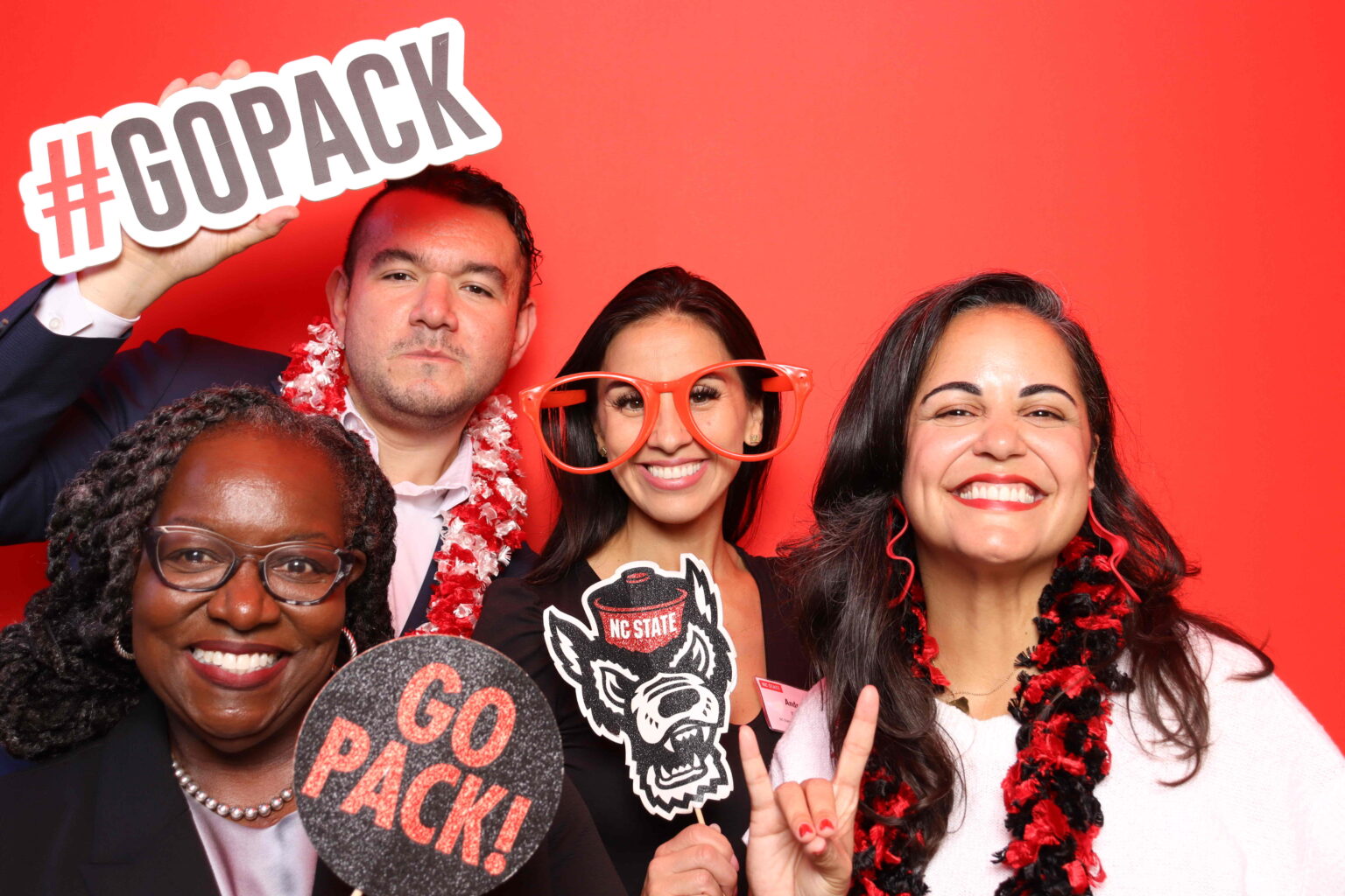 A group of NC State alumni pose and hold up festive signs in a photo booth for Red and White Week.